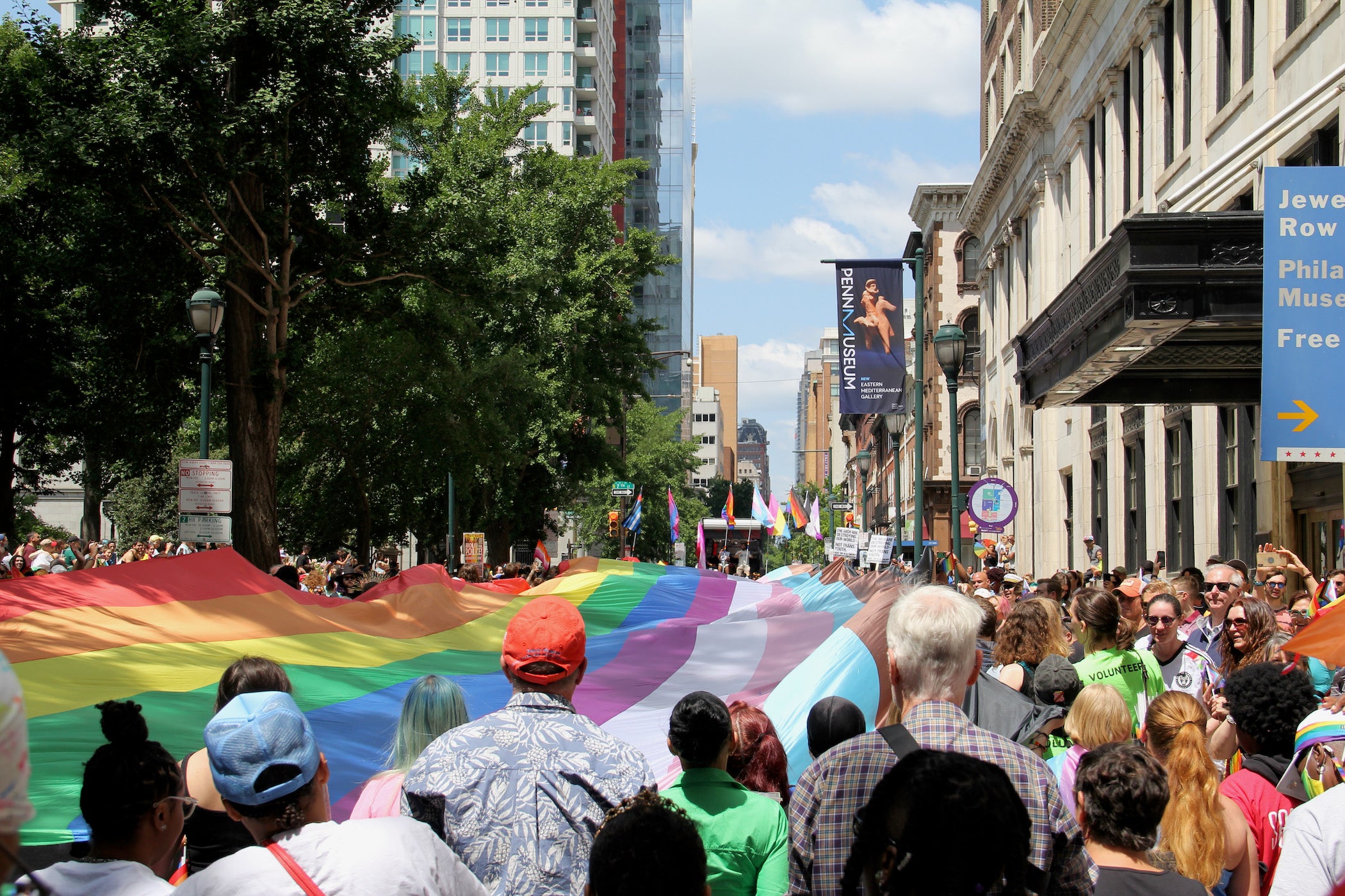 A large LGBTQ flag is carried through the streets by many people.