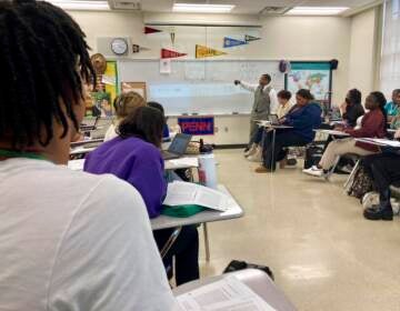 Students look towards the front of the room where their teacher is gesturing towards something written on the whiteboard.