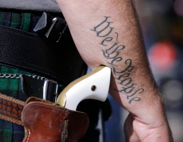 A close-up of someone's gun on a holster. On their arm is a tattoo that reads 