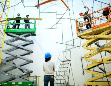 Artist Michelle Lopez wearing a construction helmet observing people on lifts installing her sculptures