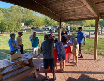 A man talks in front of a group of people standing under an open pavilion outdoors.