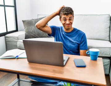 A young man sitting on the floor using a laptop with a hand on his head looking confused and uncertain.