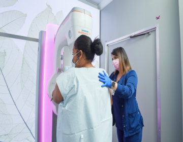 A woman steps towards a mammogram machine, guided by a health care professional.